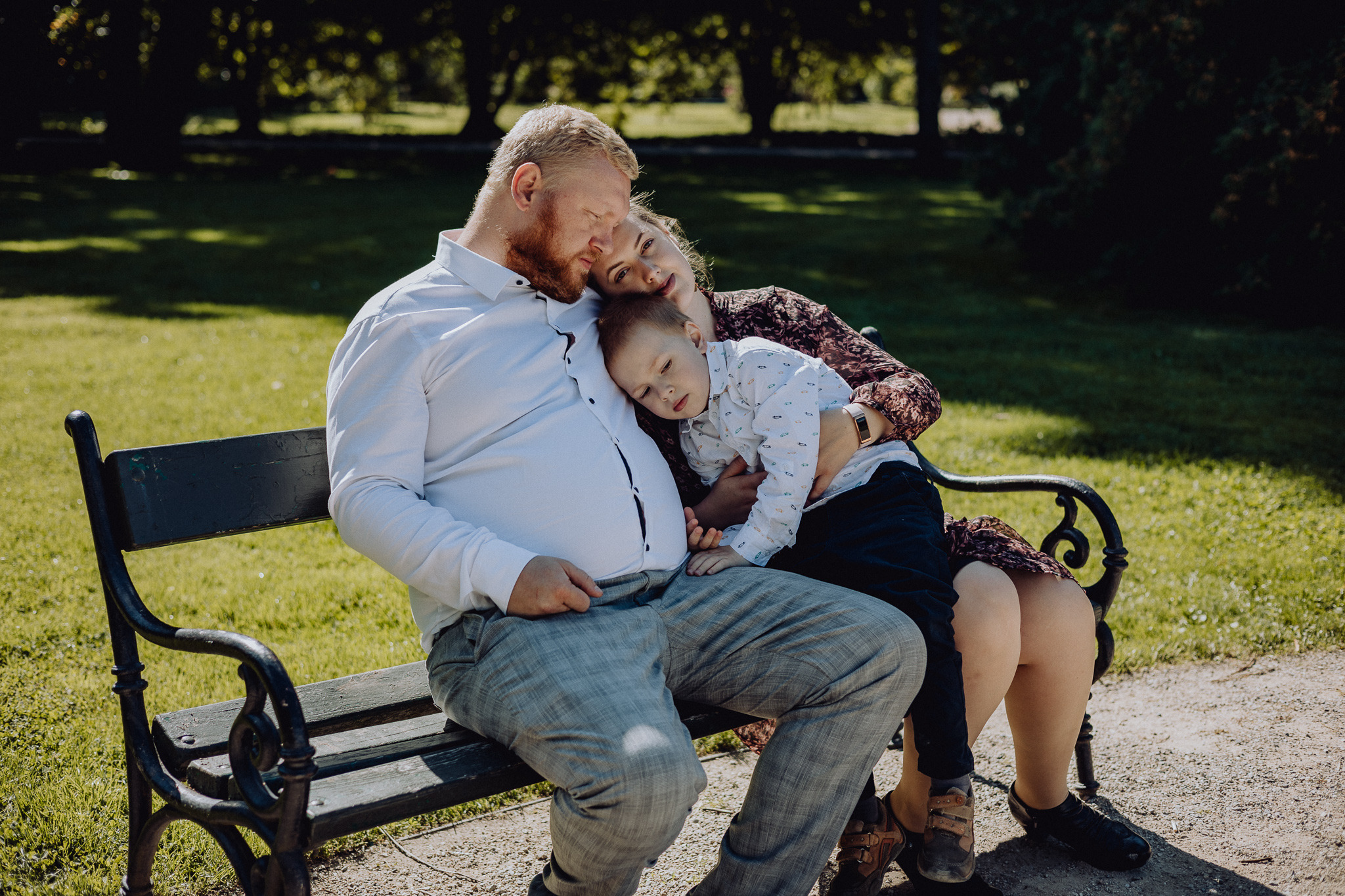 Rodzina siedząca na ławce w parku: ojciec z rudą brodą, matka z kwiatowym wzorem na sukience i syn w koszuli w autka, opierający się o ojca.