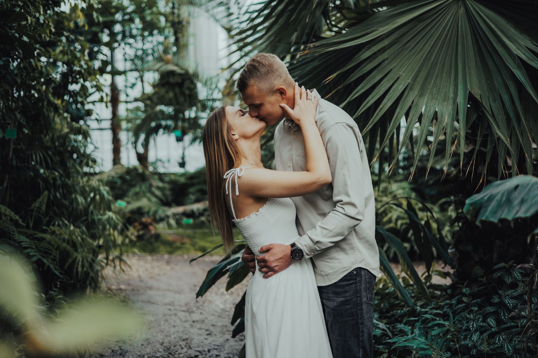 Zakochana para w białej sukience i koszuli całuje się w otoczeniu zieleni egzotycznej szklarni, mężczyzna ma na ręku zegarek.