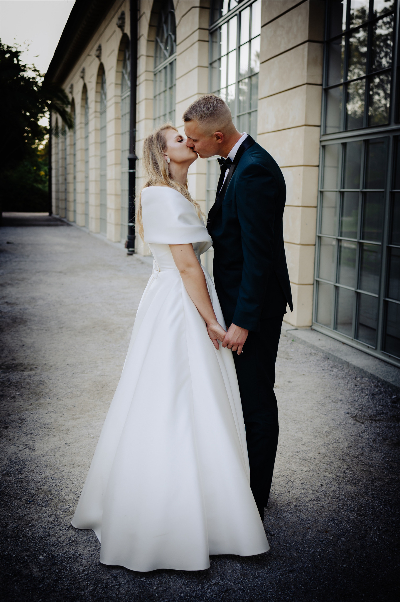 Para młoda w pocałunku na tle klasycznej architektury z dużymi oknami, panna młoda w prostej białej sukni z krótkim rękawem, pan młody w ciemnozielonym smokingu z czarną muszką.