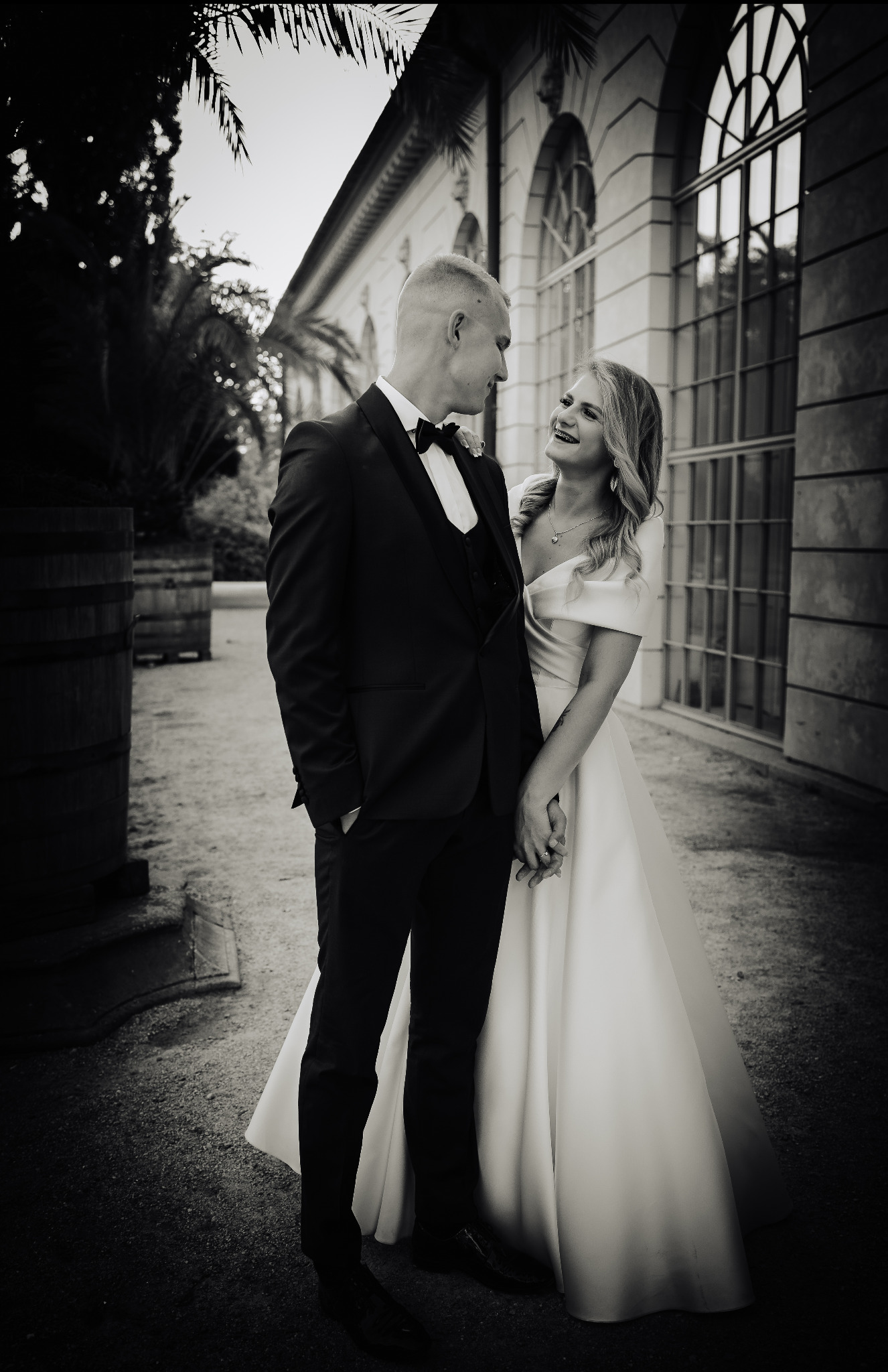 Czarno-białe zdjęcie uśmiechniętej panny młodej w satynowej sukni i pana młodego w smokingu, trzymających się za ręce na tle budynku z dużymi oknami i palmami.