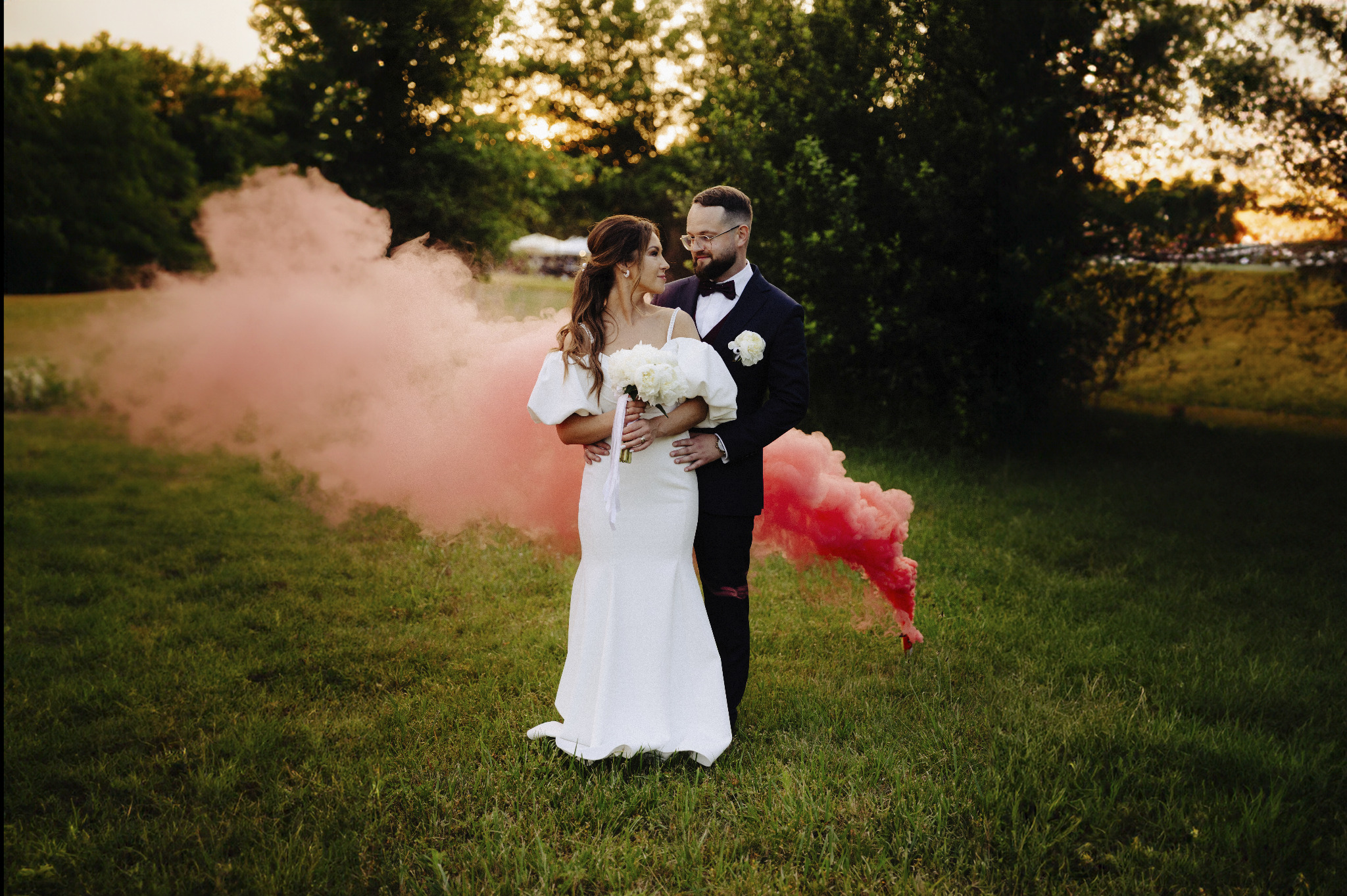 Para młoda w objęciach na łące, otoczona różową mgłą z dymnych świec. Panna młoda w białej sukni, pan młody w garniturze. Romantyczna sesja plenerowa.