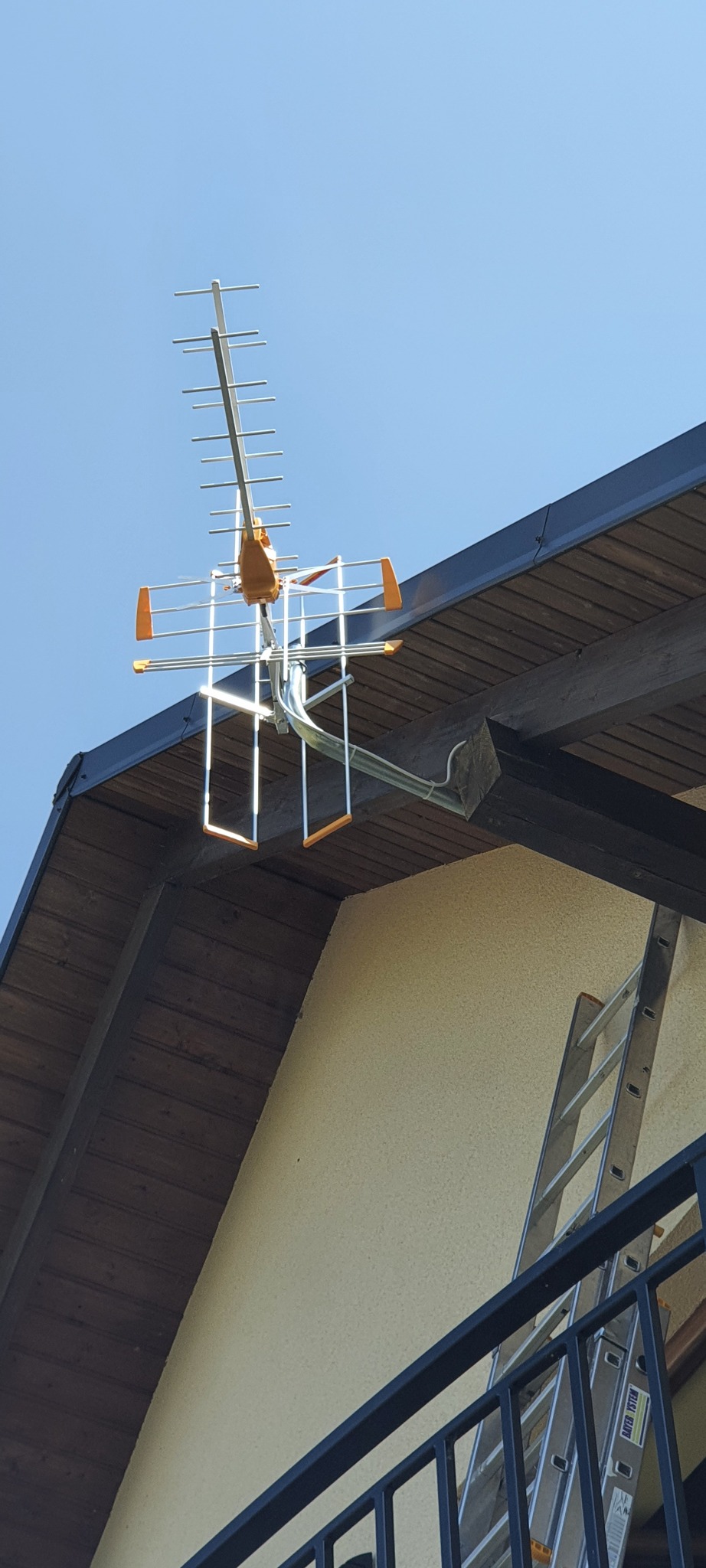 Antena telewizyjna zamontowana na drewnianym dachu budynku z beżową elewacją, widoczna drabina oparta o ścianę.