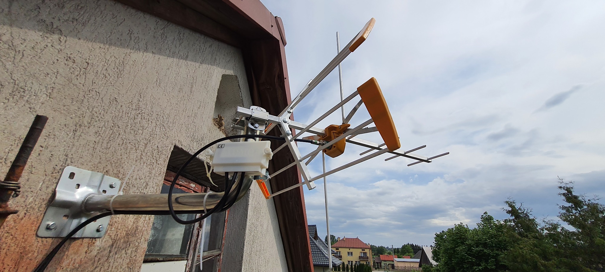 Zewnętrzna antena telewizyjna Yagi-Uda z żółtymi elementami, zamocowana do ściany budynku z widocznymi przewodami i puszką połączeniową, na tle pochmurnego nieba i zabudowań.
