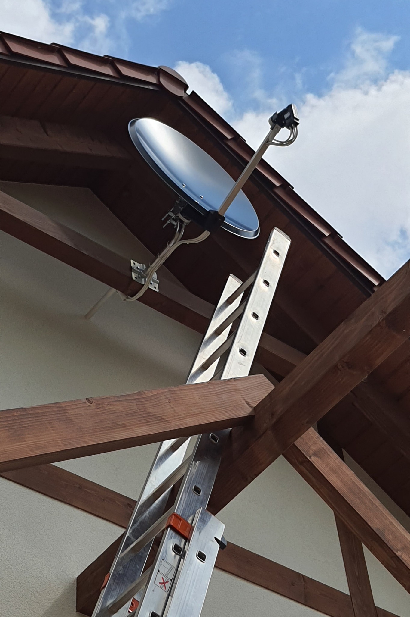 Błyszcząca antena satelitarna zamontowana na brązowym dachu, z drabiny opierającej się o konstrukcję dachu, widok z dołu na błękitne niebo z chmurami.