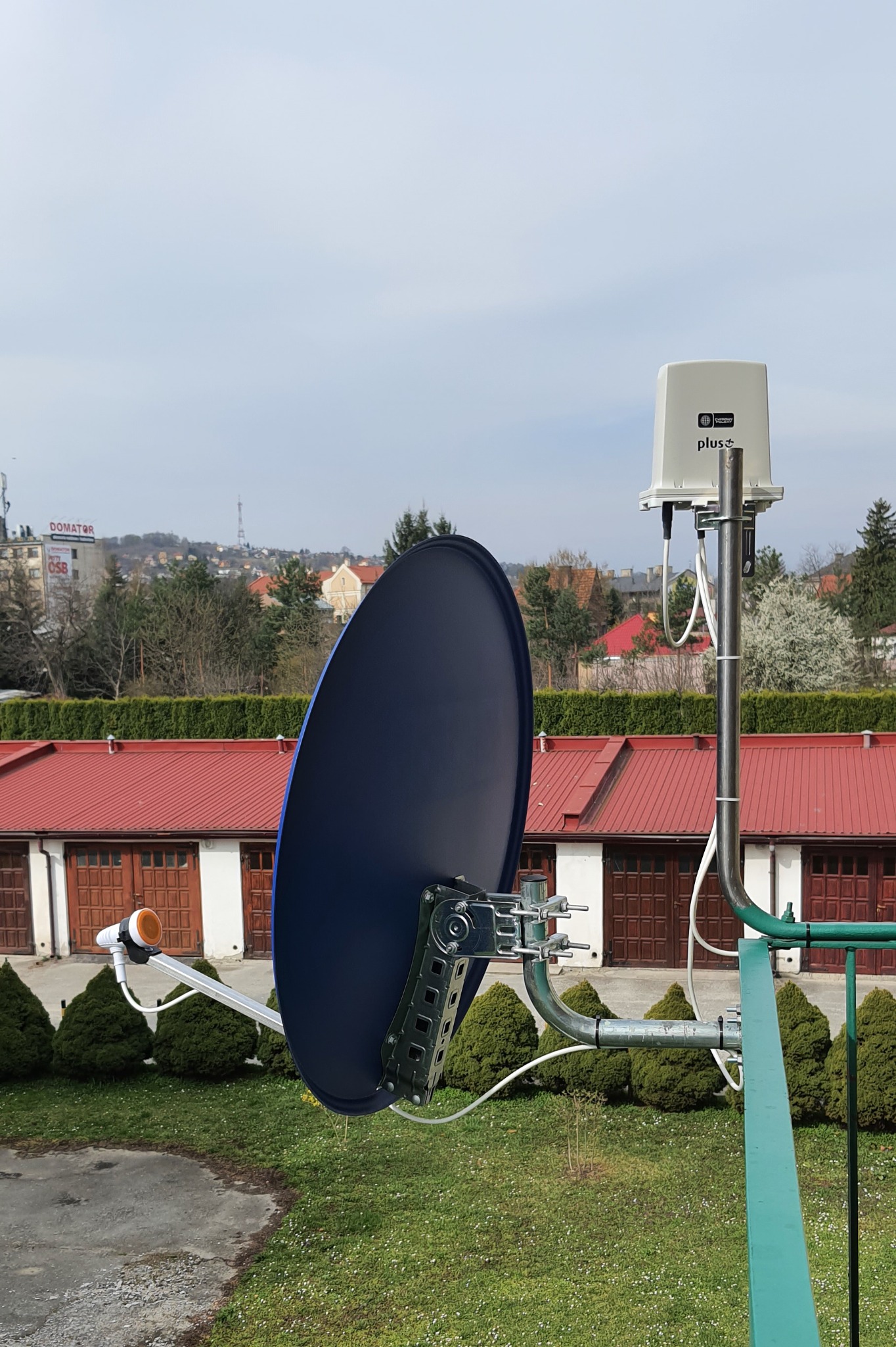 Niebieska antena satelitarna zamontowana na metalowej konstrukcji na tle zabudowań z czerwonymi dachami i zieleni, widoczny także biały odbiornik z logo 'plus+'.