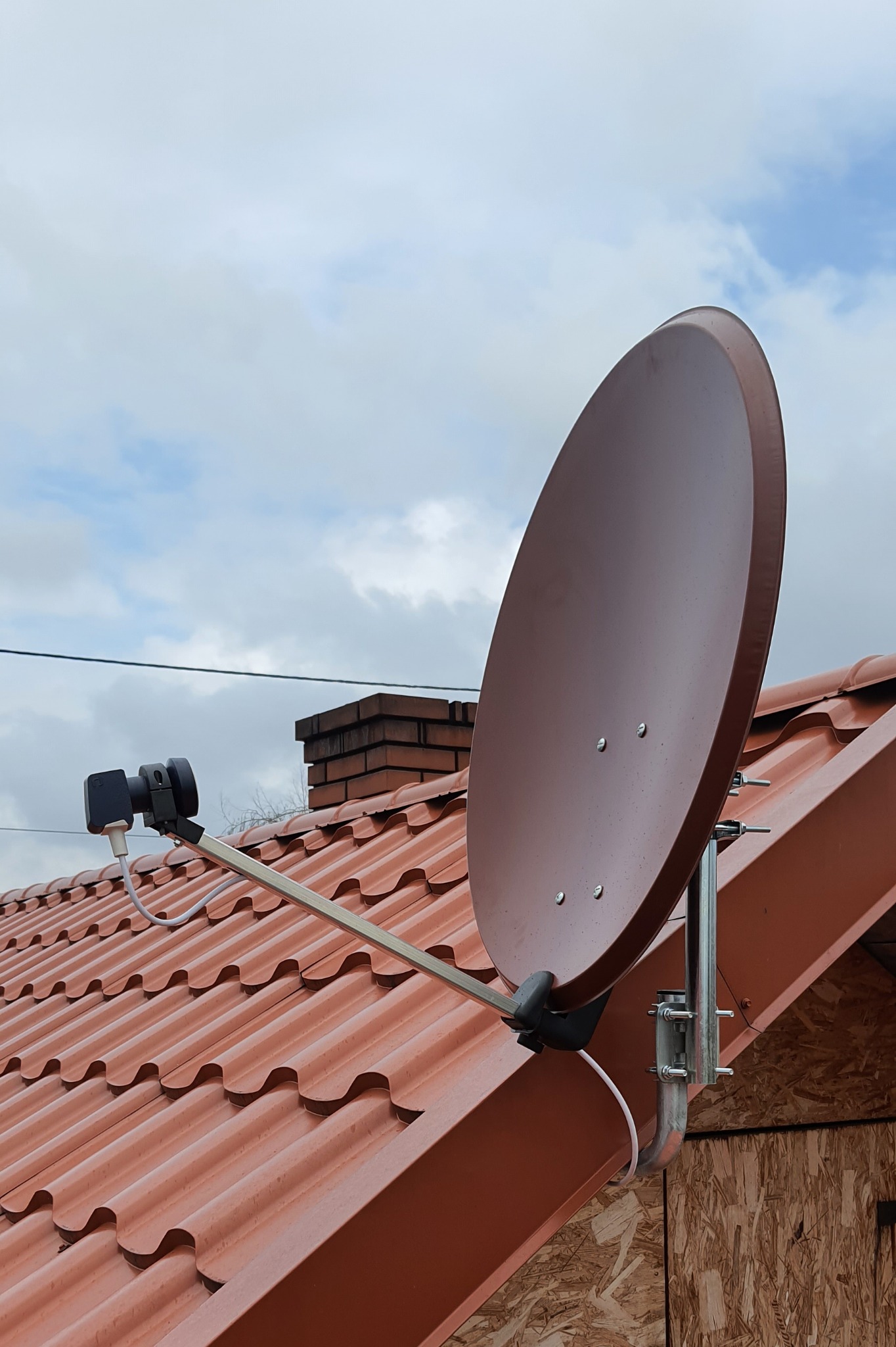 Brązowa antena satelitarna zamontowana na dachu z czerwonej dachówki, widoczny przewód antenowy i komin w tle, pochmurne niebo.