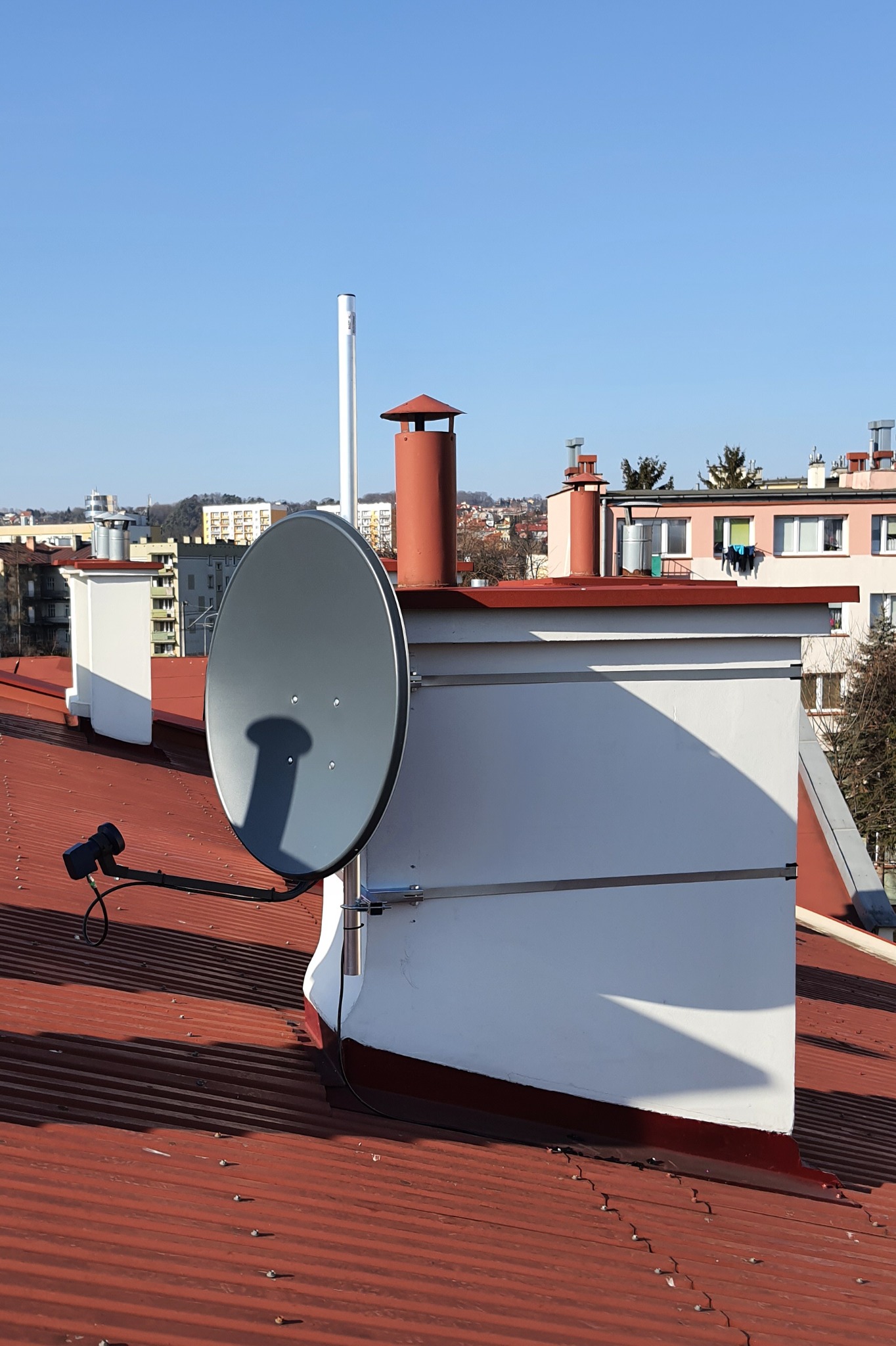 Szara antena satelitarna zamontowana na dachu budynku z czerwonej blachy, widok na miasto w tle, słoneczny dzień.
