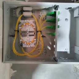 Wnętrze skrzynki telekomunikacyjnej z rozdzielnicą światłowodową, widoczne żółte kable światłowodowe i zielone złącza, zamontowana na ścianie z widocznymi śladami tynku.