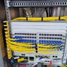 Szafa telekomunikacyjna z patch panelem, światłowodami i urządzeniami sieciowymi Nokia, widoczne kable światłowodowe w kolorze żółtym i niebieskim, zasilacze Flatpack i przełączniki z portami RJ45.