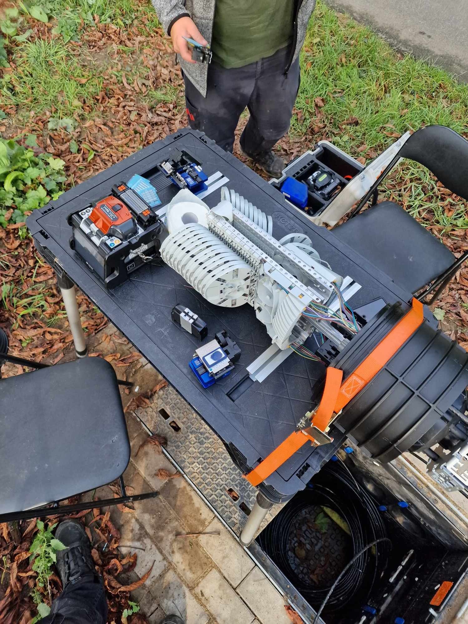 Technik łączy światłowód na przenośnym stole roboczym na zewnątrz, widoczne narzędzia i elementy złączek.