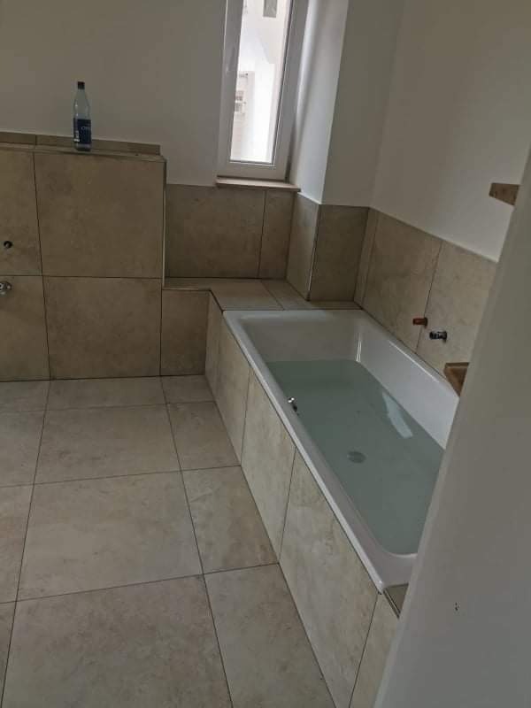 Beżowe płytki w łazience, wanna wypełniona wodą, butelka wody mineralnej na obudowie wanny.