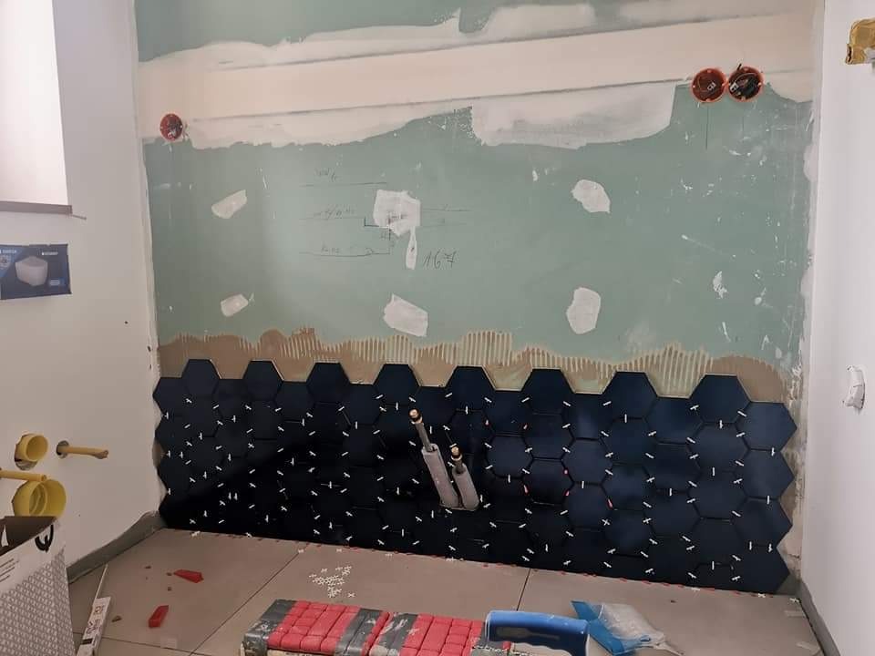 Montaż ciemnych, heksagonalnych płytek na ścianie w łazience, widoczne rurki i poziomica, białe krzyżyki dystansowe między płytkami, surowa ściana przygotowana pod dalsze prace wykończeniowe.