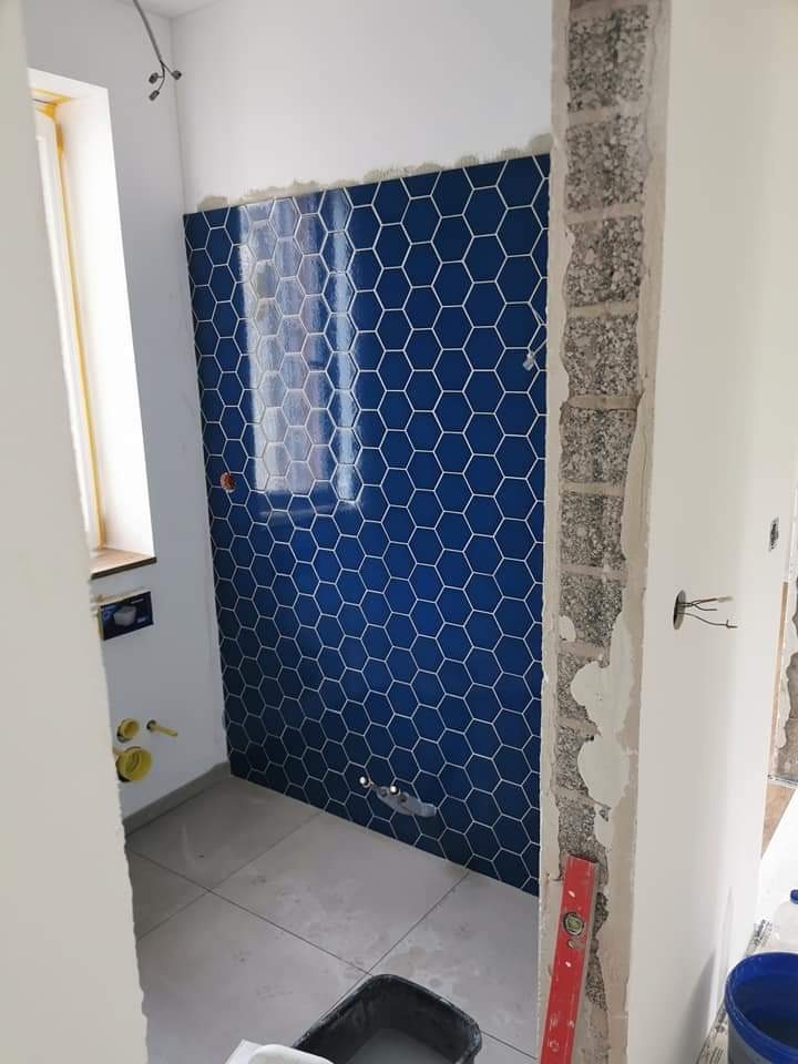 Niebieskie heksagonalne płytki częściowo ułożone na ścianie łazienki w trakcie remontu, widoczne przygotowane instalacje, poziomica przy ościeżnicy i wiadro z zaprawą na podłodze.