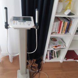 Dietetyk Kliniczny mgr Ewelina Duda - Profesjonalny analizator składu ciała z uchwytami na dłonie, na tle białej ściany i regału z książkami o tematyce zdrowotnej i żywieniowej.