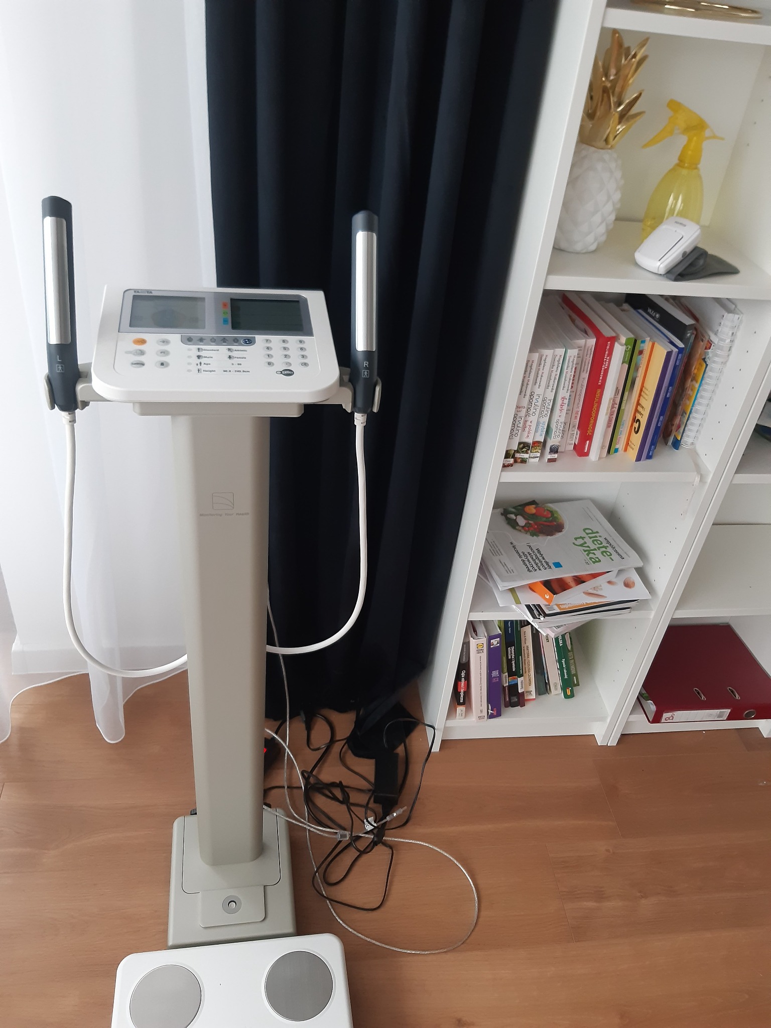 Profesjonalny analizator składu ciała z uchwytami na dłonie, na tle białej ściany i regału z książkami o tematyce zdrowotnej i żywieniowej.