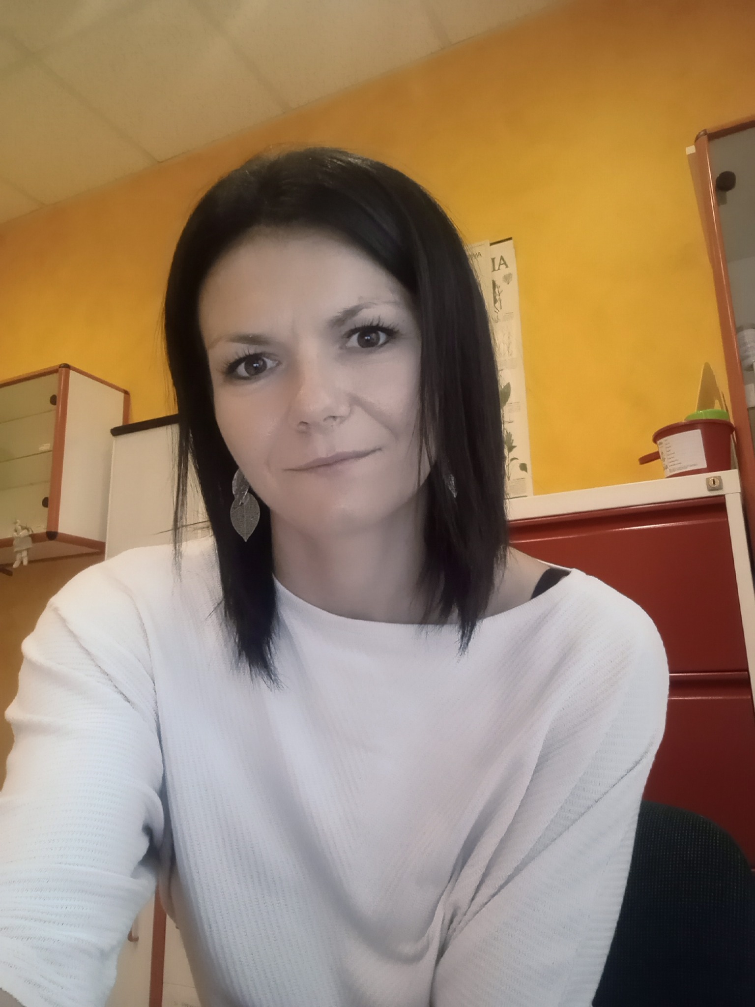 Portret kobiety z ciemnymi włosami, w białej bluzce, z kolczykami w kształcie liści, na tle żółtej ściany i czerwonej komody.