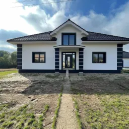 Nowoczesny dom jednorodzinny z białymi ścianami, czarną stolarką okienną i dachem z ciemnej dachówki, widziany z perspektywy ścieżki prowadzącej do wejścia, na tle błękitnego nieba z chmurami.