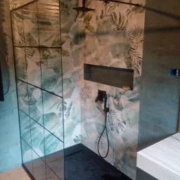 Kabina prysznicowa typu walk-in z czarną armaturą, heksagonalnymi płytkami w odcieniach zieleni z motywem roślinnym i wnęką na kosmetyki z gąbką w środku.