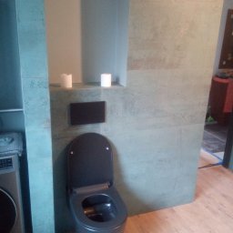 Solid Krzysztof Stachański - Nowoczesna toaleta w kolorze grafitowym z deską wolnoopadającą, zamontowana na ścianie wyłożonej płytkami imitującymi beton, z widocznym fragmentem pralki i drewnopodobną podłogą.