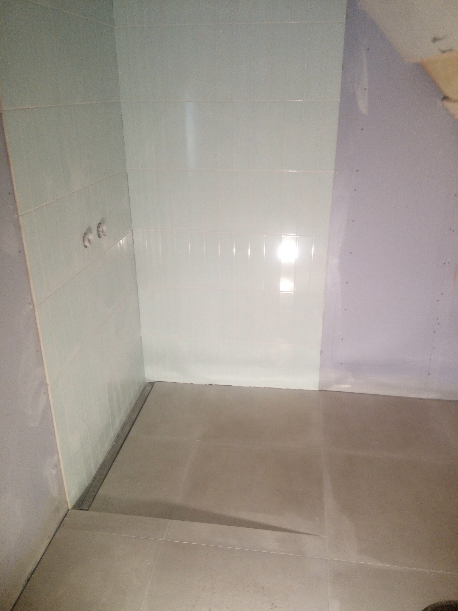 Nierównomiernie oświetlona, częściowo wykończona łazienka z jasnymi płytkami ściennymi i szarymi podłogowymi, widoczne przyłącza wodne i surowa ściana z płyt kartonowo-gipsowych.