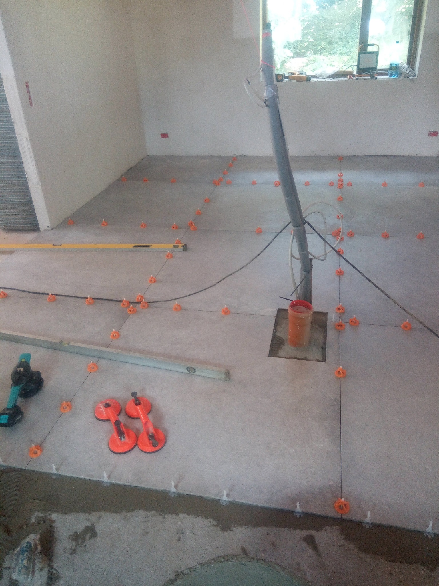 Układanie płytek podłogowych: widoczne poziomice, przyssawki do płytek, sznurki wyznaczające linie, pomarańczowe krzyżyki dystansowe i białe kliny poziomujące na szarej podłodze w trakcie prac...