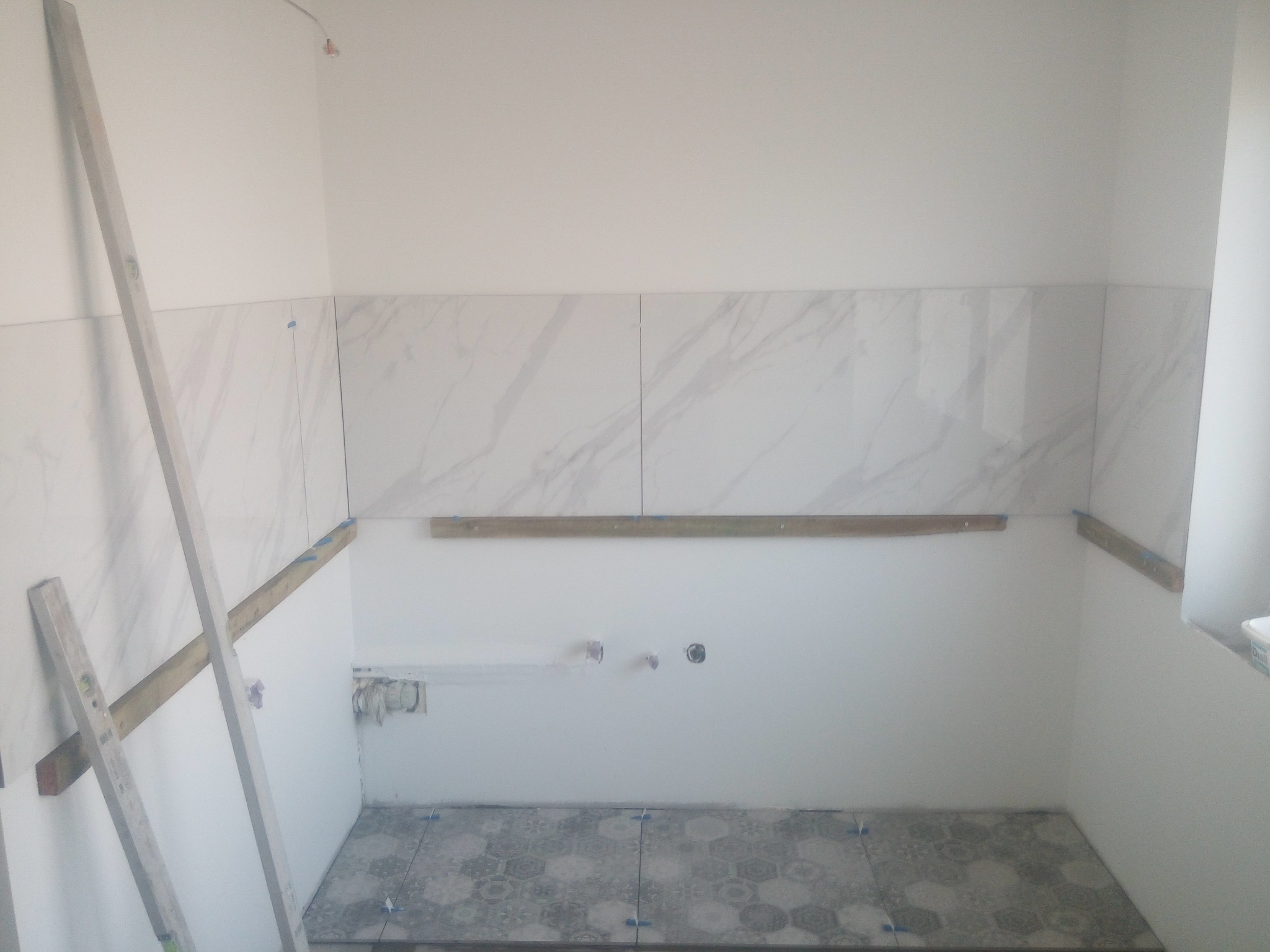 Wnętrze łazienki w trakcie układania płytek: marmurowe płytki ścienne do połowy wysokości, szare płytki podłogowe z geometrycznym wzorem, drewniane listwy pomocnicze, widoczne instalacje...