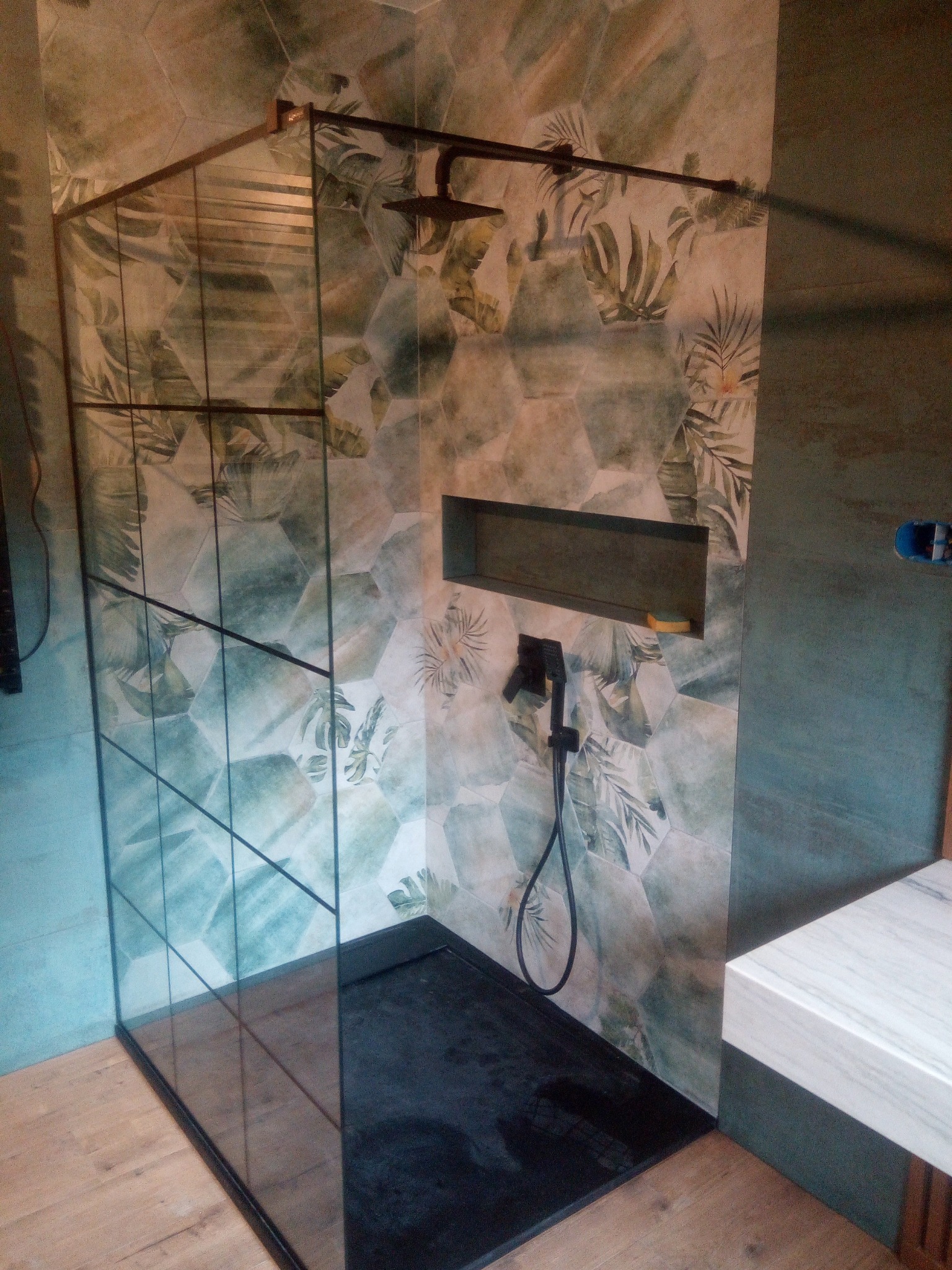 Kabina prysznicowa typu walk-in z czarną armaturą, heksagonalnymi płytkami w odcieniach zieleni z motywem roślinnym i wnęką na kosmetyki z gąbką w środku.