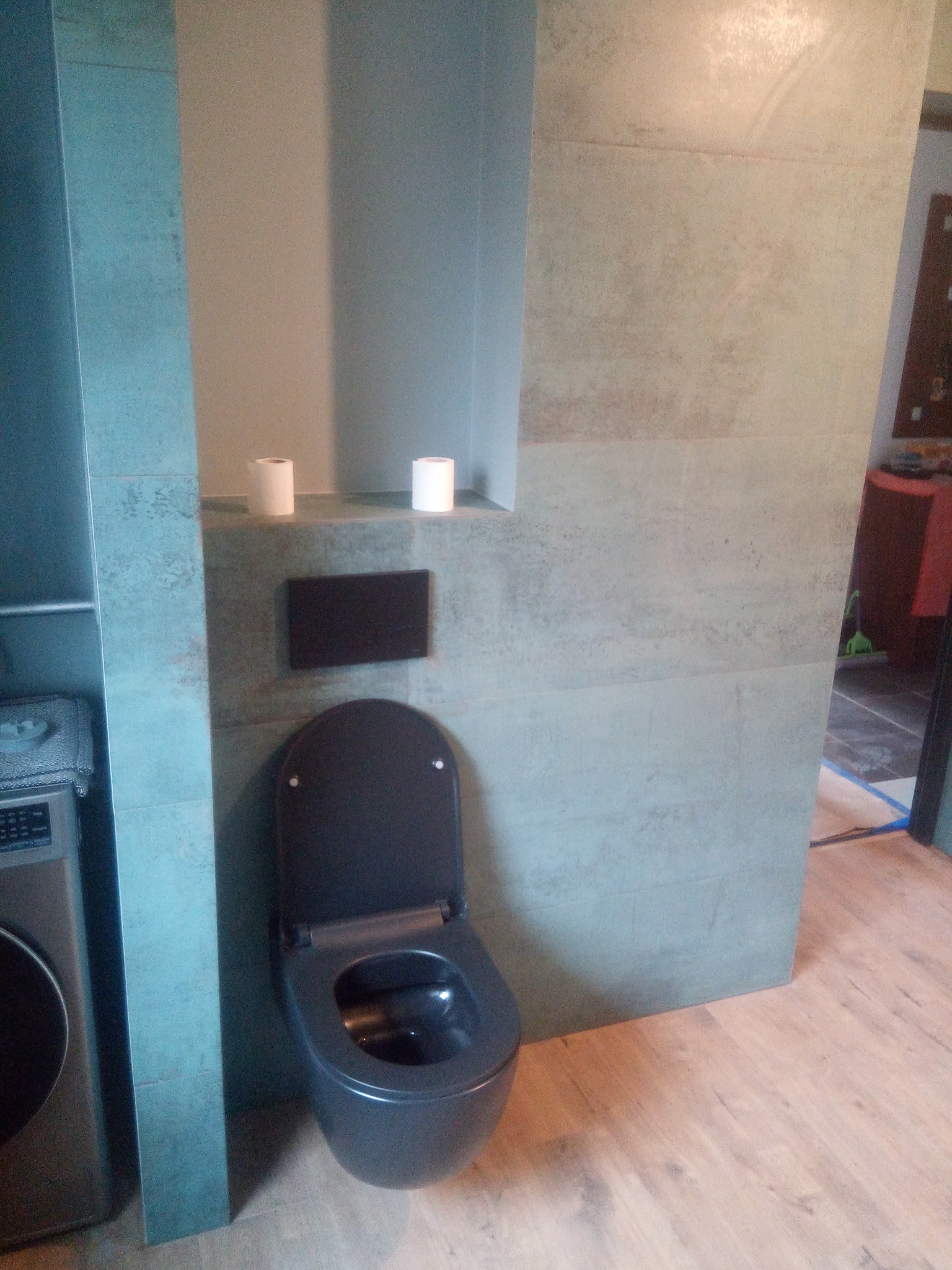 Nowoczesna toaleta w kolorze grafitowym z deską wolnoopadającą, zamontowana na ścianie wyłożonej płytkami imitującymi beton, z widocznym fragmentem pralki i drewnopodobną podłogą.