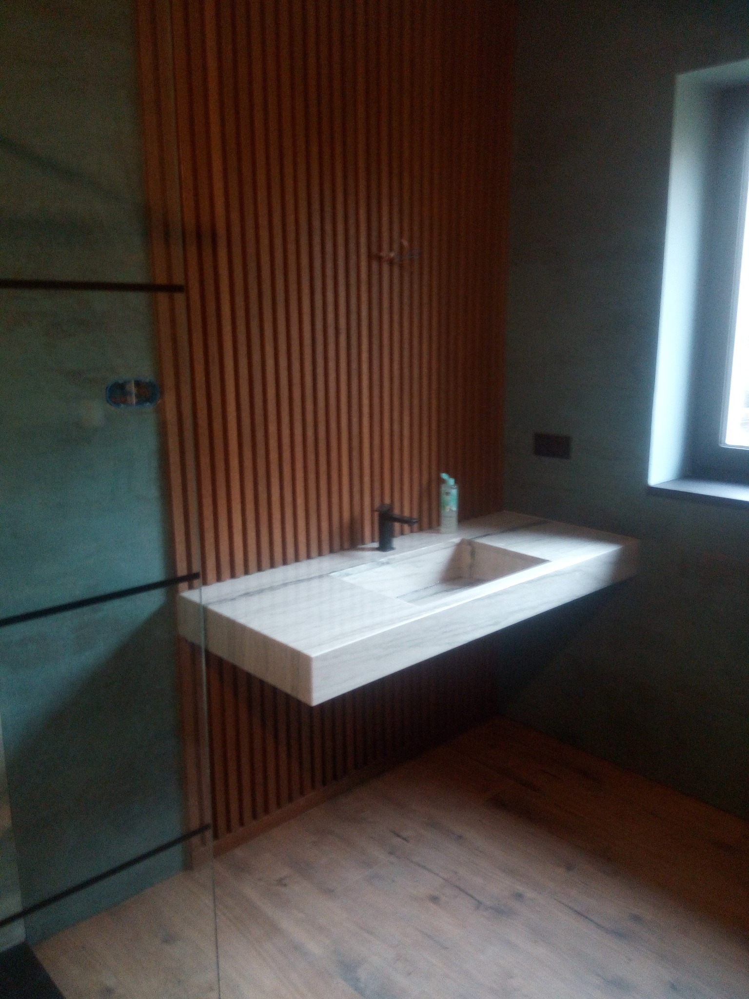 Nowoczesna łazienka z umywalką z jasnego marmuru zawieszoną na tle ściany z drewnianych listew, widoczny fragment kabiny prysznicowej z grafitowymi elementami oraz podłoga z paneli imitujących drewno.