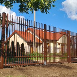 Klanse sp. z o.o. - Brązowa metalowa brama wjazdowa i ogrodzenie panelowe z pionowych prętów, widok na dom z czerwoną dachówką w słoneczny dzień.
