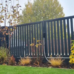 Nowoczesna, antracytowa brama wjazdowa z pionowych profili, z automatyką i skrzynką na listy, otoczona ozdobną roślinnością jesienną.