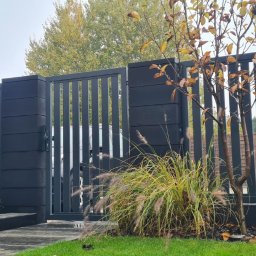 Klanse sp. z o.o. - Nowoczesne, antracytowe ogrodzenie panelowe z furtką i słupkami z bloczków betonowych, otoczone ozdobną trawą i młodymi drzewkami z jesiennymi liśćmi.