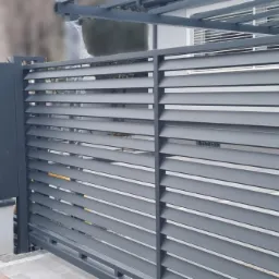 Nowoczesne, szare ogrodzenie posesji z poziomych paneli, z wbudowaną skrzynką na listy, widoczne z zewnątrz.