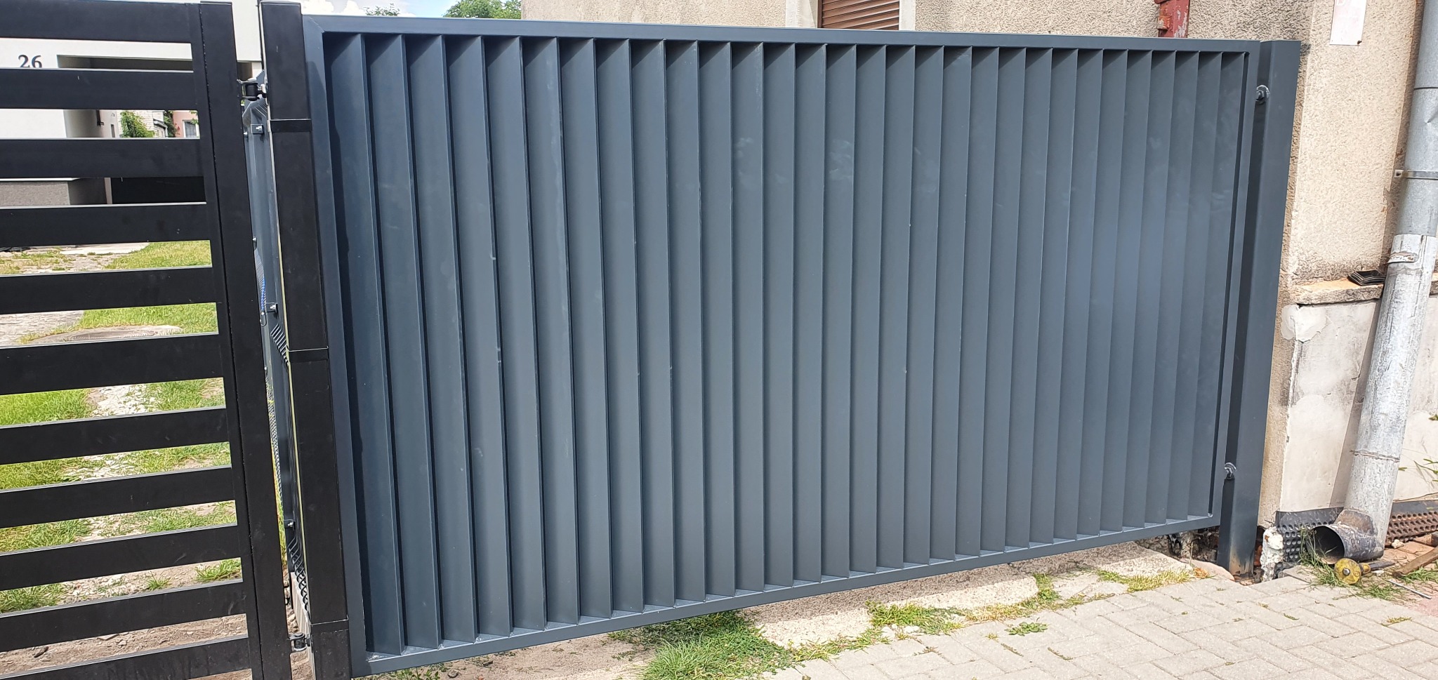Nowoczesna, przesuwna brama wjazdowa w kolorze grafitowym, z pionowymi panelami, zamontowana przy budynku z widoczną rynną spustową.