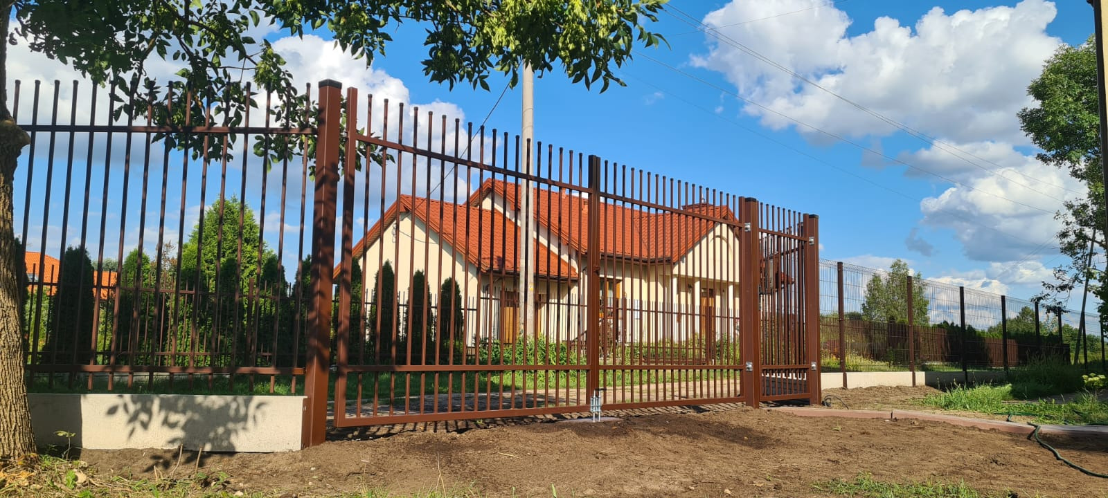 Brązowa metalowa brama wjazdowa i ogrodzenie panelowe z pionowych prętów, widok na dom z czerwoną dachówką w słoneczny dzień.