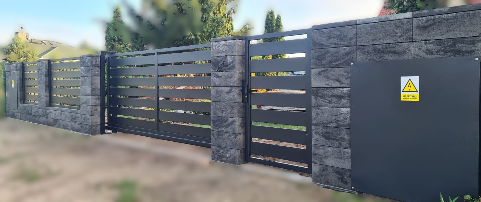 Nowoczesna brama wjazdowa przesuwna z poziomymi panelami, osadzona na słupkach z szarej kostki imitującej kamień, obok skrzynka elektryczna z żółtą naklejką ostrzegawczą.