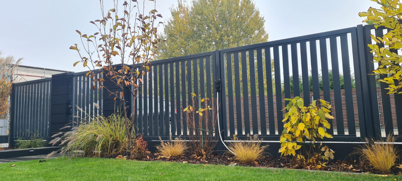 Nowoczesna, antracytowa brama wjazdowa z pionowych profili, z automatyką i skrzynką na listy, otoczona ozdobną roślinnością jesienną.