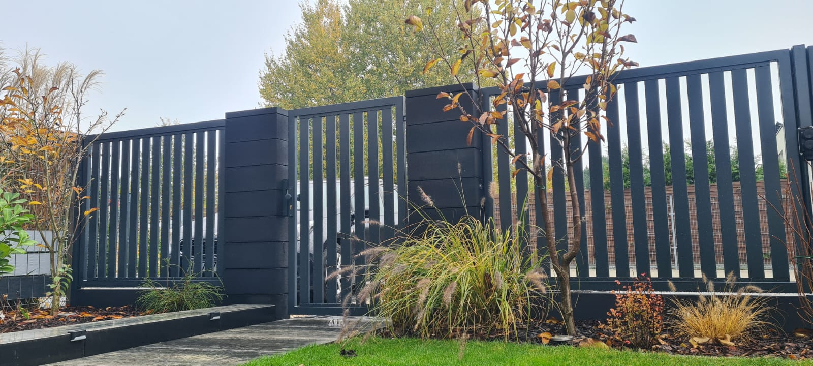 Nowoczesne, antracytowe ogrodzenie panelowe z furtką i słupkami z bloczków betonowych, otoczone ozdobną trawą i młodymi drzewkami z jesiennymi liśćmi.