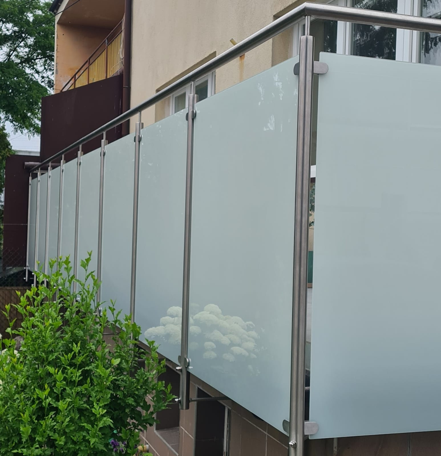 Nowoczesna balustrada balkonowa ze stali nierdzewnej i mlecznego szkła, widok z dołu, z zielenią w tle.