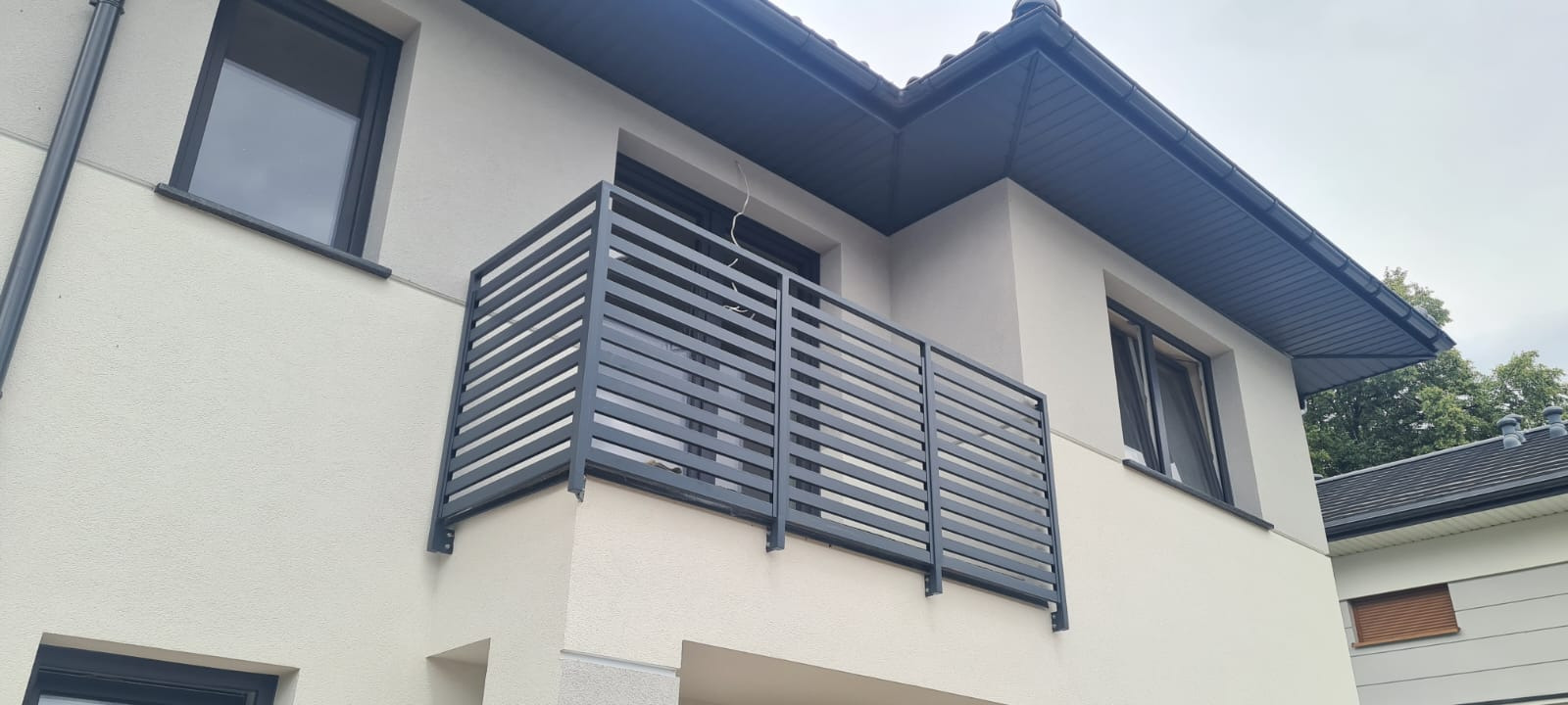 Nowoczesna, grafitowa balustrada balkonowa z poziomych profili, zamontowana na jasnej elewacji budynku z grafitową podbitką dachową i oknami w tym samym kolorze.
