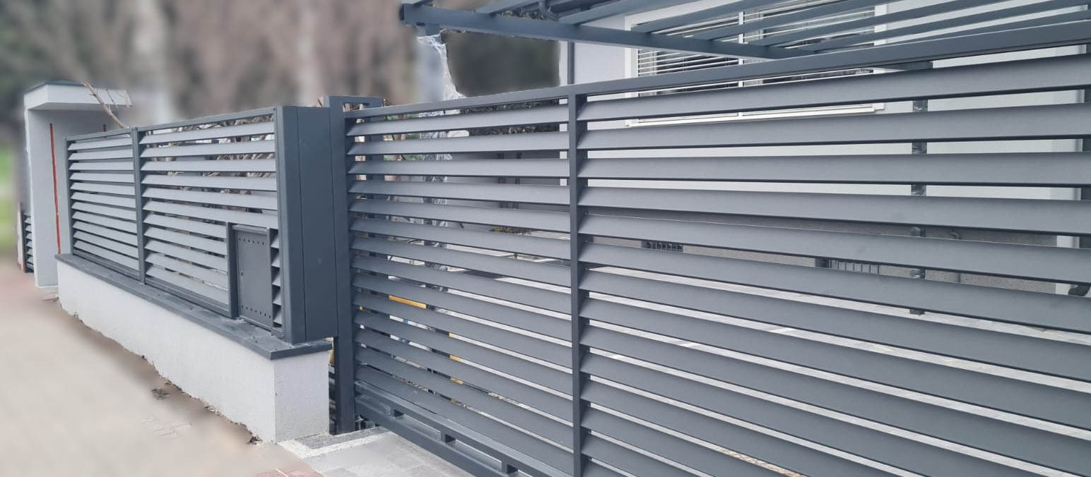 Nowoczesne, szare ogrodzenie posesji z poziomych paneli, z wbudowaną skrzynką na listy, widoczne z zewnątrz.