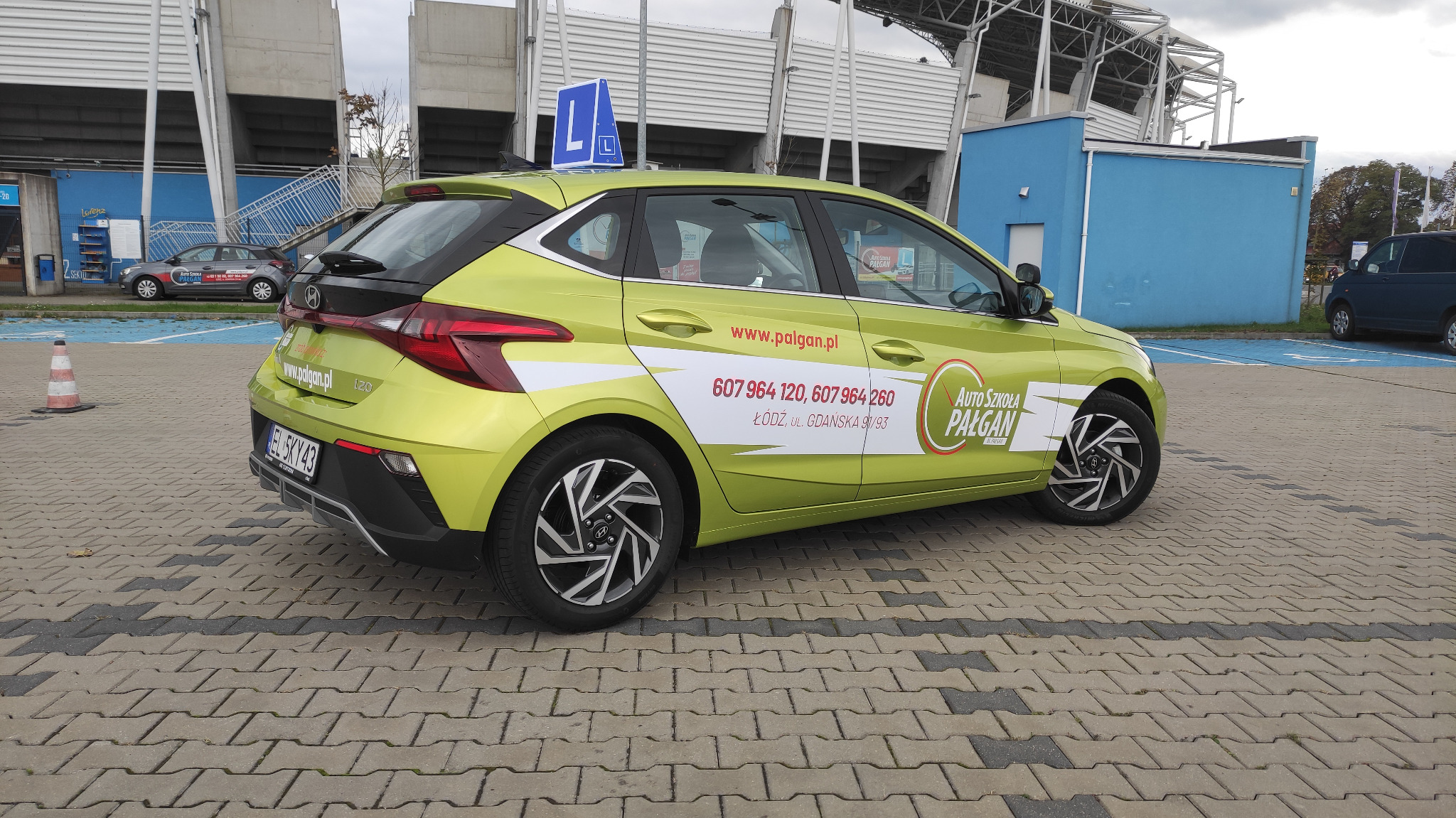 Limonkowy samochód z oznaczeniami Auto Szkoły Pałgan zaparkowany na parkingu z kostki brukowej przed stadionem, widoczna tabliczka z literą L na dachu.