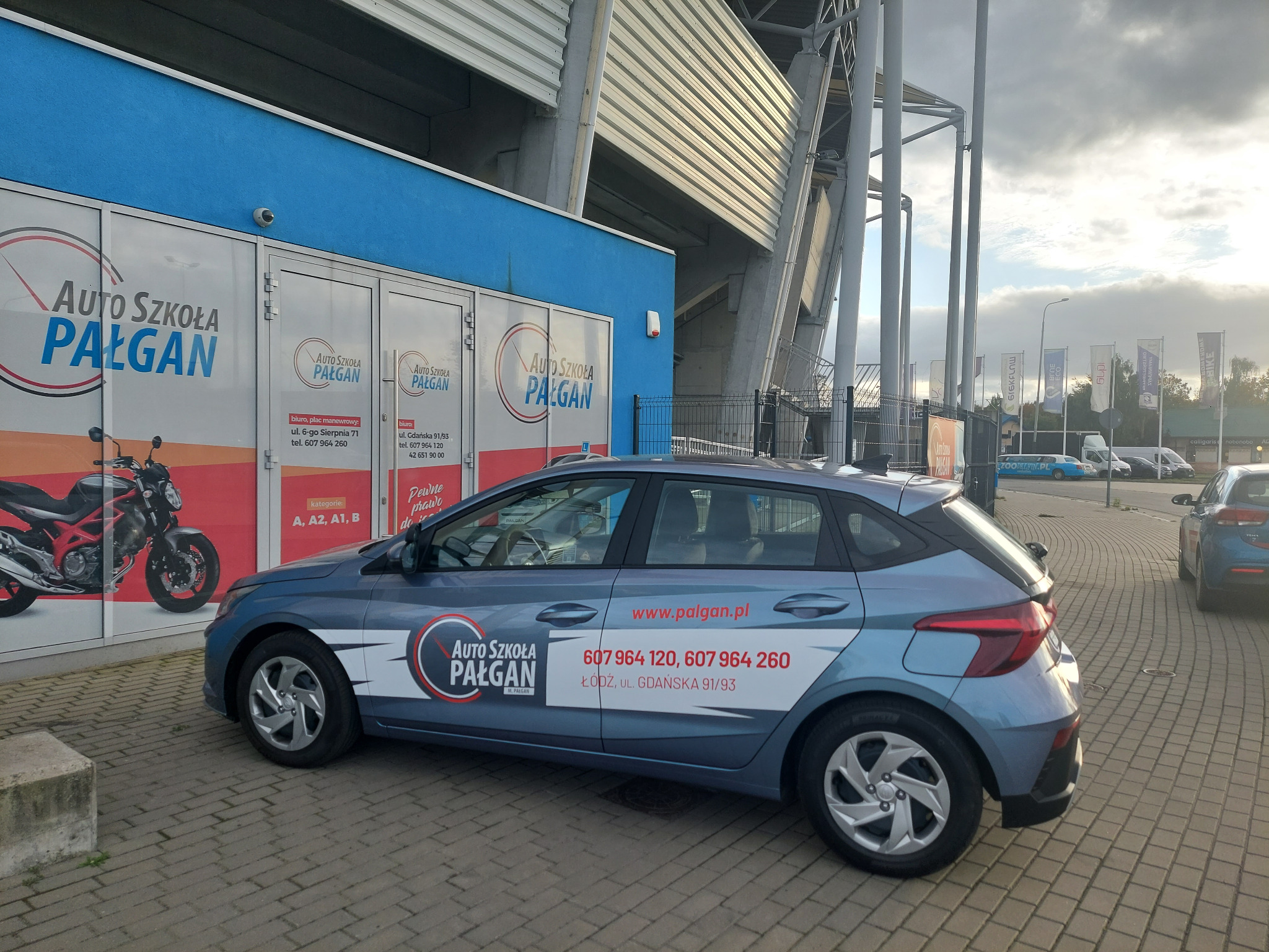 Niebieski samochód z logo i numerem telefonu szkoły nauki jazdy Pałgan, zaparkowany na kostce brukowej przed budynkiem z reklamą szkoły i motocyklem na witrynie, w tle widoczny stadion.