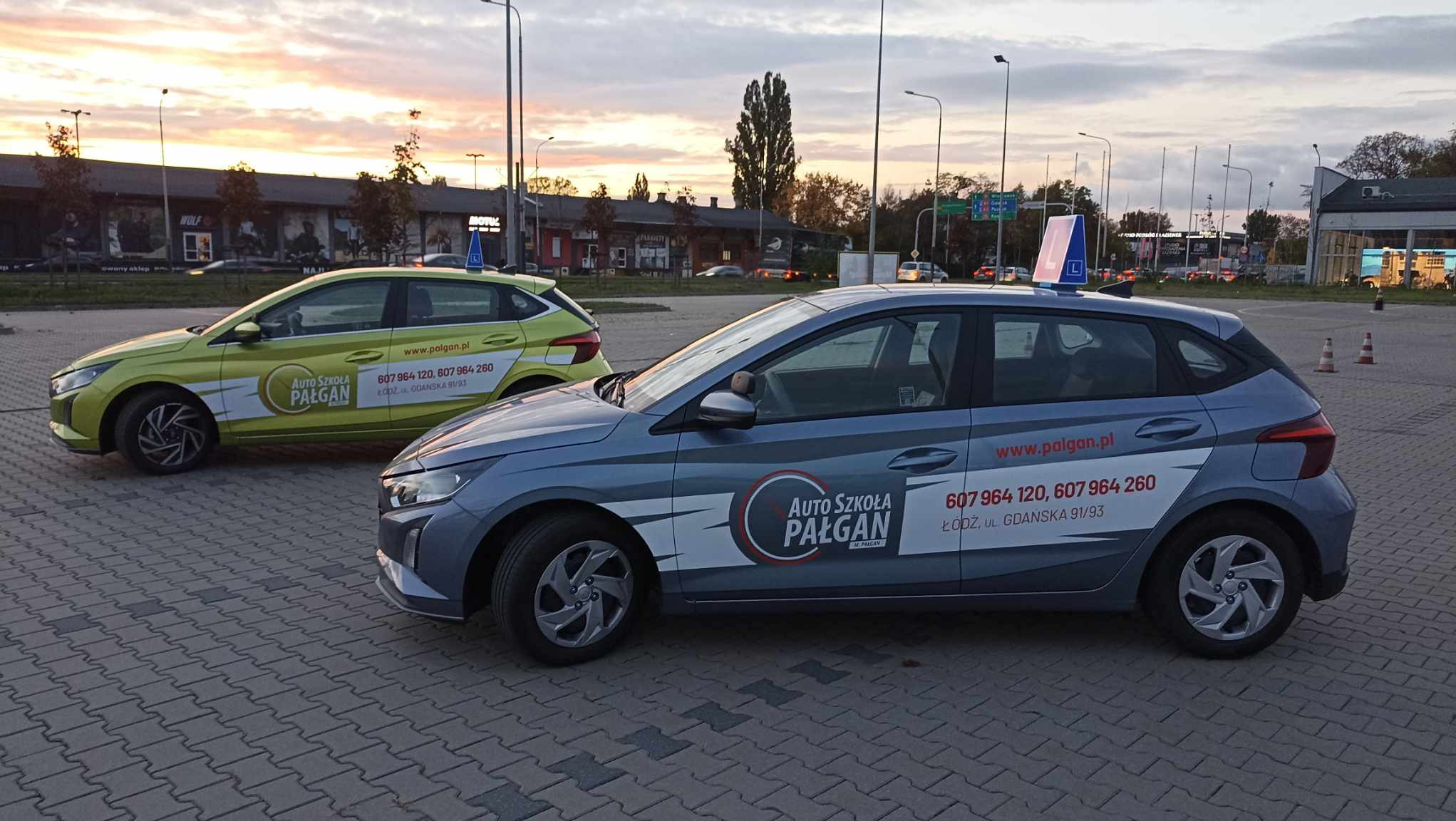 Dwa samochody z oznaczeniami szkoły nauki jazdy z Łodzi, zaparkowane na placu manewrowym. Na dachu niebieskiego auta widoczna tabliczka z literą 'L'.