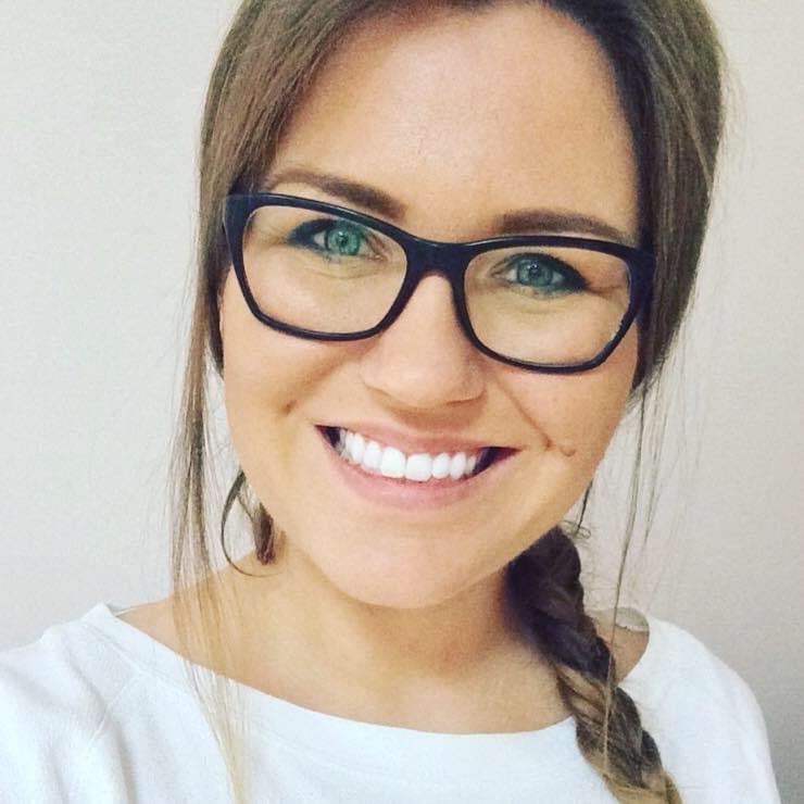 Uśmiechnięta kobieta w okularach z ciemnymi oprawkami i warkoczem, prezentująca białe zęby na tle jasnej ściany.