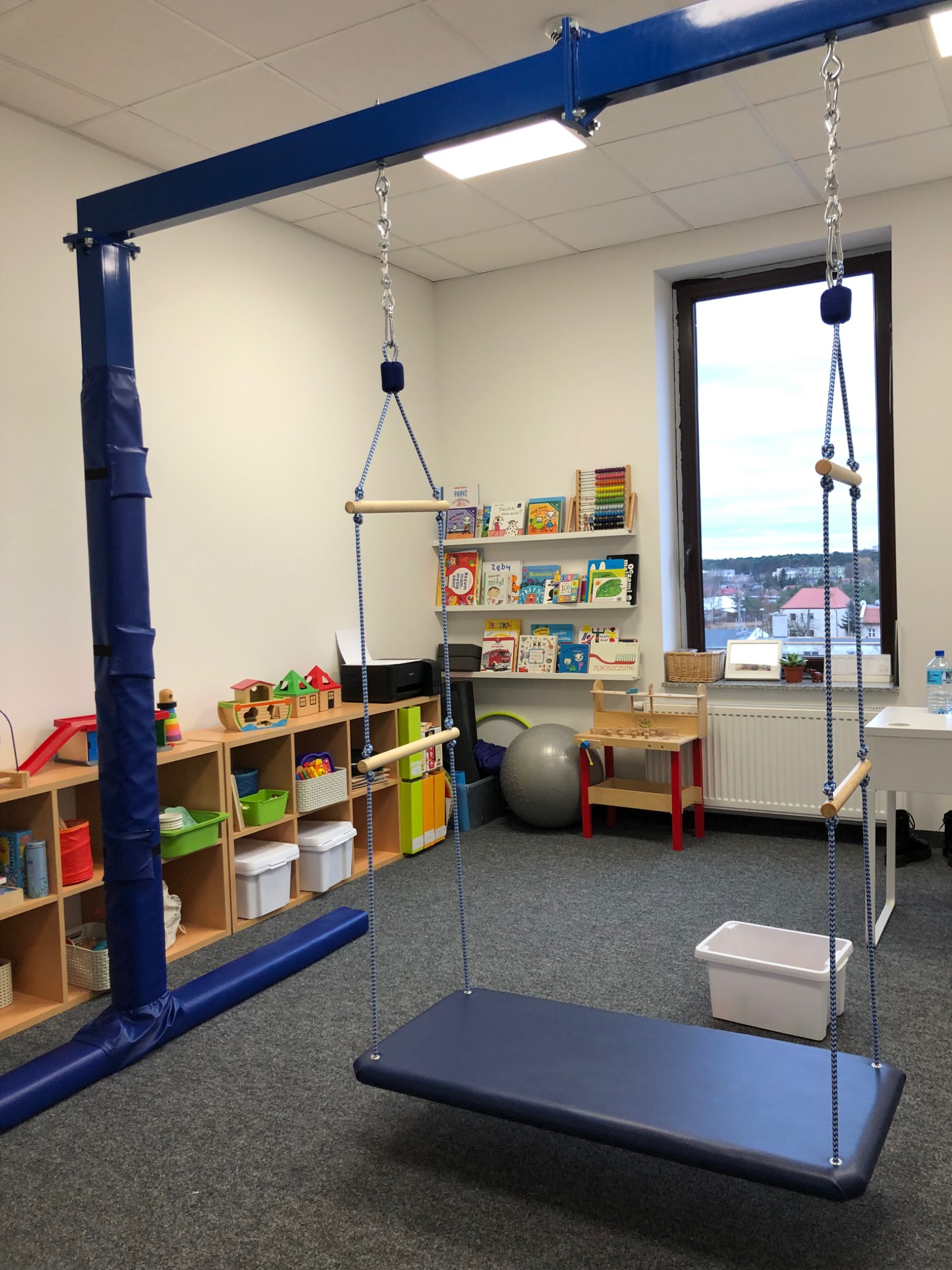 Sala terapii integracji sensorycznej z huśtawką podwieszaną, drabinką sznurkową, regałami z zabawkami i książkami, widok z okna na miasto.