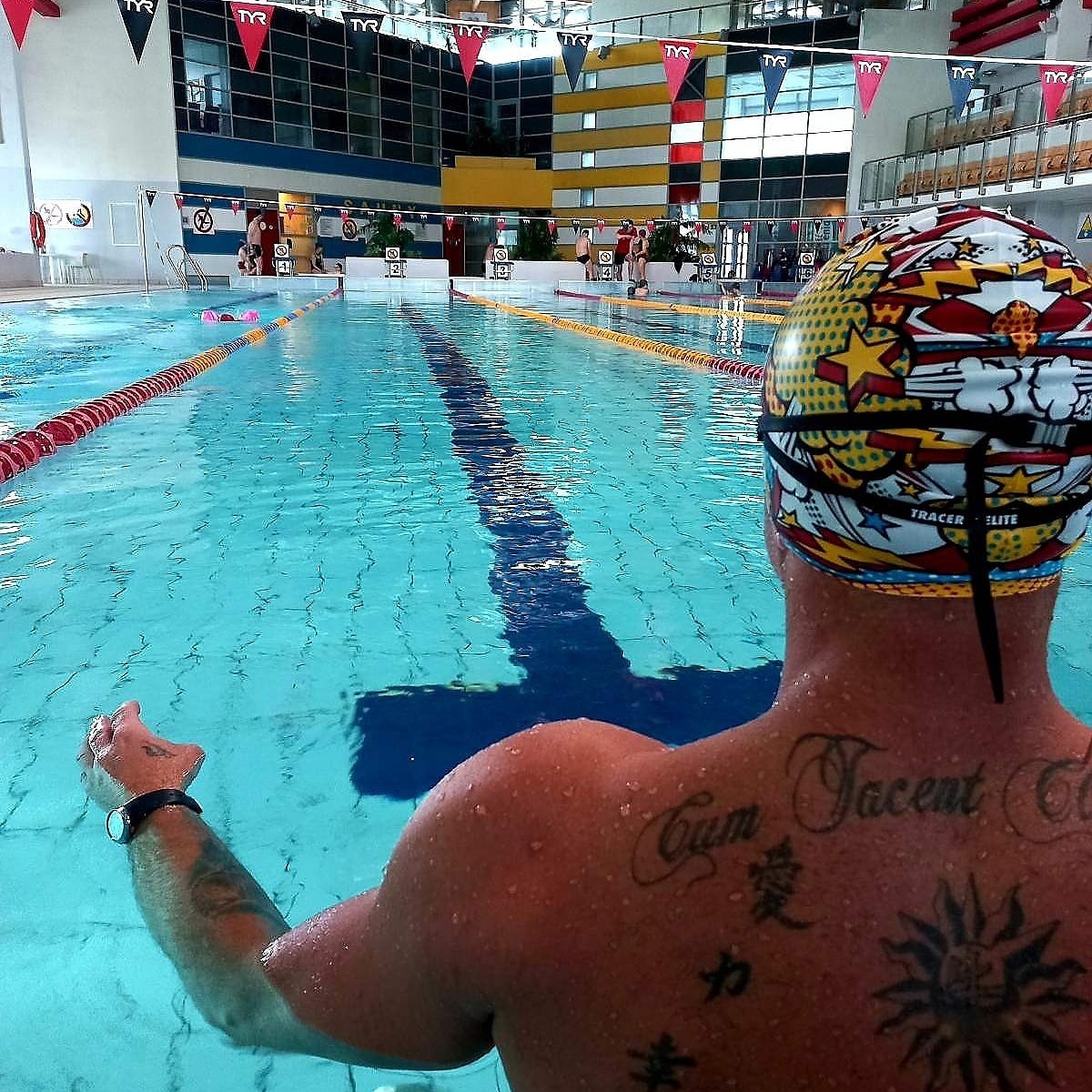 Mężczyzna w basenie, widziany od tyłu, w czepku pływackim z komiksowym wzorem i tatuażami na plecach, woda sięga do ramion, w tle tor pływacki z flagami.