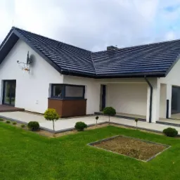 Nowoczesny dom jednorodzinny z białą elewacją, czarnym dachem i stolarką okienną, z anteną satelitarną na ścianie i zagospodarowanym terenem wokół.