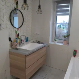 Nowoczesna łazienka z drewnianą szafką pod umywalką, białymi płytkami, okrągłym lustrem, wzorzystą tapetą i wiszącymi lampami w stylu industrialnym.