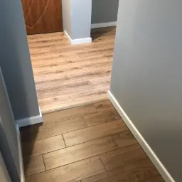 Połączenie dwóch rodzajów podłóg: ciemnobrązowych płytek imitujących drewno i jasnych paneli laminowanych w korytarzu z szarą ścianą i białymi listwami przypodłogowymi.